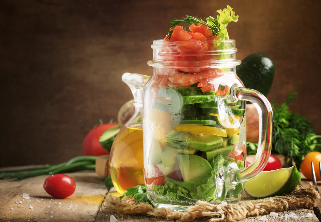Salad with vegetables, avocado, shrimp and trout, source of omega-3, served in glass jar, wood kitchen table, selective focus