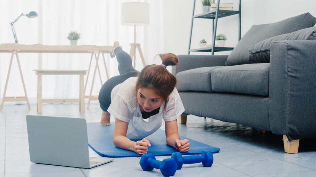 Young lady in sportswear exercises doing working out planks with a leg outstretched and using laptop to watching yoga video tutorial at home. Distant training with personal trainer, social distance.